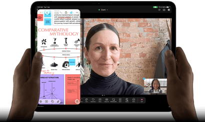iPad Pro, front exterior, landscape orientation, space black colour, rounded corners, black display bezel, hands holding left and right sides, display showing a FaceTime conversation being live translated, and an educational app showing a presentation of Comparative Mythology with text, diagrams and handwritten notes